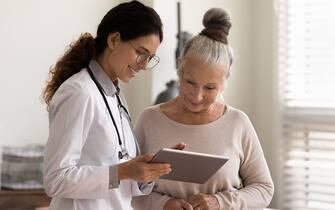 Happy female doctor and senior patient look at tablet screen discuss treatment or therapy on gadget. Smiling woman nurse and mature client use pad device discussing results in modern hospital.