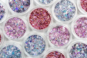 Blue, pink and red glitter in jars on white background. Shiny holographic elements, sparkle. Beautiful shimmer for manicure, makeup, nail extension, design. Cosmetic products. Trendy delicate colors.