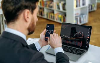 Exchange market. Smart man investor and crypto trader, using laptop and cellphone, analyzes charts of trading in stock market and digital cryptocurrency exchange,conducts analysis,trading crypto coins