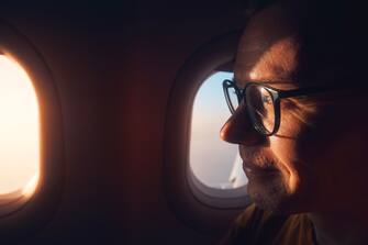 Portrait of man with eyeglasses in airplane. Passenger looking through window during flight at sunset.