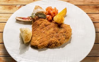 Milanese cutlet, veal cutlet with bone breaded and fried in butter. In white dish with potatoes and tomatoes on wooden table