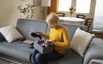 A woman, sitting on a couch in her living room is examines the contents of the opened box, holding her new purchase of boots in her hands