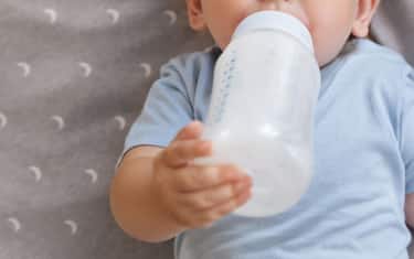 Upper view of adorable baby boy holding bottle with formula lying in crib, enjoying milk or tasty healthy nutritious baby formula. Bottle-feeding concept. Childcare