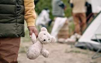 Hand of homeless child holding white teddybear while standing against refugee camp