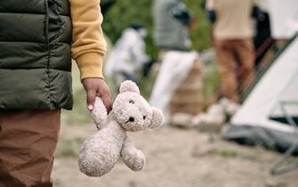 Hand of homeless child holding white teddybear while standing against refugee camp