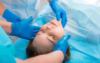 Young woman lies on surgical table before surgery. Surgeon patient portrait on table for rhinoplasty operations with doctors hands in gloves. Surgery