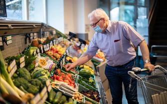 Senior man with face mask buying vegetables in a local supermarket. Elderly man doing home shopping in grocery store with a shopping cart.