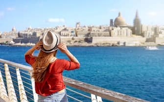 Tourism in Europe. Traveler girl holding hat walking along Malta promenade with Valletta cityscape on the background.