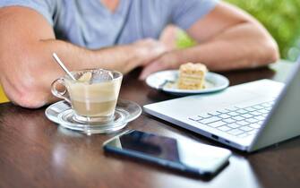 Young guy freelancer working on laptop in cafe. on the table there is cup of cappuccino coffee, piece of cake on saucer, smartphone