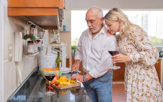 Domestic scene of a man cooking next to a woman drinking wine