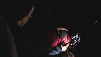 Ukraine: Grandmother holds radio in hands sitting in darkness blackout during russian terroristic attacks. Senior woman in winter clothes in darkness