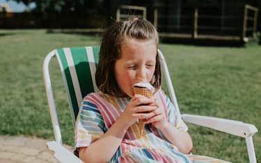 A little girl cools down on a hot day, sprawled out on a green folding chair in a domestic garden, wearing a summer dress, and eating an ice cream.