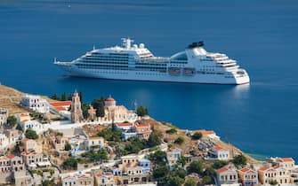 Hilltop houses and Greek Orthodox church dwarfed by cruise ship anchored offshore, Gialos (aka Yialos), Symi (aka Simi), Rhodes, Dodecanese Islands, South Aegean, Greece, Europe.