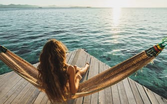 Amazing vacations in Asia. Leisure and relaxation of young beautiful woman in white swimsuit. Hammock on lounge terrace of hotel with over-reef ocean view. Wanderlust travel. Beautiful and natural morning or sunset light.