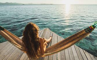Amazing vacations in Asia. Leisure and relaxation of young beautiful woman in white swimsuit. Hammock on lounge terrace of hotel with over-reef ocean view. Wanderlust travel. Beautiful and natural morning or sunset light.