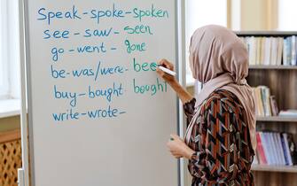 Unrecognizable woman wearing hijab standing at whiteboard doing task with irregular verbs during English language for immigrants lesson