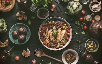 Grilled chicken breast with Mediterranean sauce in cooking pan and ingredients:  tomatoes, artichokes, olives, sun-dried tomatoes,figs, anchovies, fresh seasoning herbs and spices on dark rustic table with vintage kitchen utensils , top view.