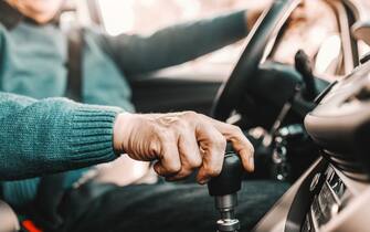 Close up of senior man holding one hand on gearshift and other on steering wheel while sitting in his car. Selective focus on hand.