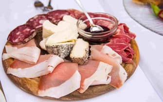 A mixed platter of ready-sliced meats, Lardo Piemontese pepato, Prosciutto Crudo di Cuneo, salamino crudo cacciatore, and Langa cow cheeses with a bowl of strawberry grape jam on a restaurant table in La Morra, Piedmont, Italy