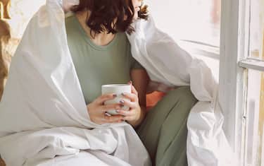 Relaxed positive young brunette woman in homewear sitting on top of unmade bed with cup of tea in her hands, slowly drinking aromatic hot beverage whi