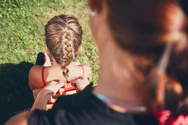 Young woman making hair braids of the little girl on the garden in the countryside.