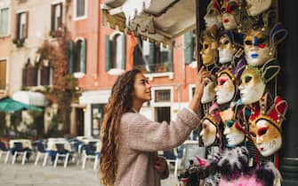 Vacations and shopping in Venice, Italy. Tourist woman want to choose and buy one. Traditional beautiful handmade carnival masks as souvenir selling on street kiosk. Giel enjoying and smiling.
