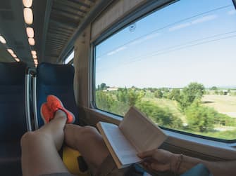 Young adult man lying on the seat of a passenger train and reading a book. Wanderlust concept