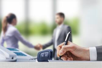 Customer and car salesman shaking hands at the car dealership, businessman signing a contract in the foreground