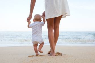 Baby boy steps with mother and baby learning to walk on sandy beach in Portugal.