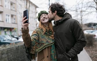 A man kissing his girlfriend on the cheek as she takes a selfie of the two of them while out for a walk in the city together.
