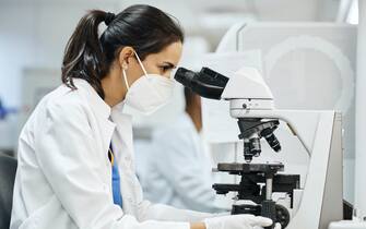 Side view of female doctor using microscope during research in laboratory. Healthcare professional is working in hospital during COVID-19. She is wearing protective face mask and lab coat.