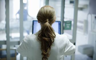 Woman doctor with long red hair using laptop in hospital, photographed from behind