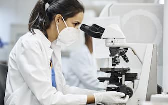 Side view of female doctor using microscope during research in laboratory. Healthcare professional is working in hospital during COVID-19. She is wearing protective face mask and lab coat.