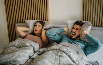 Annoyed adult woman and man in bed suffering neighbor noise at home. The couple has an unhappy facial expression closing their ears with pillows while lying in bed.