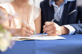 Close-up view of man and woman signing marriage certificate, she wears a vintage wedding dress and he wears a formal suit. Beginning of life as a couple, with a promise to be together forever.