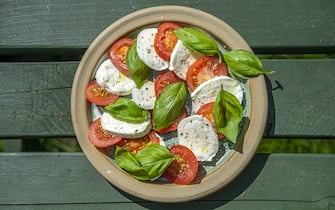 A close-up aerial view of a plate of freshly prepared Italian-style caprese salad with a green wooden table top beneath. Caprese salad is a simple Italian salad, made of sliced fresh mozzarella, tomatoes, and sweet basil, drizzled with olive oil. It features the colours of the Italian flag: green, white, and red. In Italy, it is usually served as an antipasto (starter).