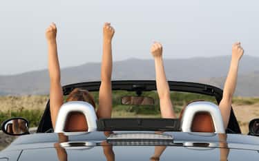 Back view portrait of two women raising arms in a convertible car enjoying views in the mountain