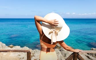 A woman in a hat and bikini stands by a wooden fence and looks at the sea.