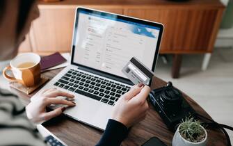 Over the shoulder view of young Asian woman shopping online for flight tickets on airline website with laptop, entering credit card details to make mobile payment at home. Camera and passport on the table. Travel planning. Booking a holiday online