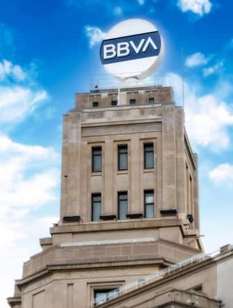 Logo or sign of the BBVA Spanish bank on top of the rectangular tower. The stone wall building is seen on an overcast weather day in the city center.