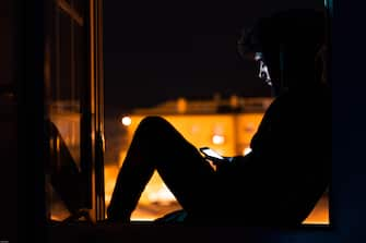 Young man with mobile phone confined to his house watching out the window at night.Patient isolated in quarantine to prevent coronavirus infection.