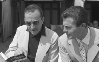 Regista e attore Italian film director Luchino Visconti, wearing a fair suit, a striped shirt and sunglasses, smoking a cigarette, holding a book about his movie 'White Nights', sitting next to actor Marcello Mastroianni, wearing a suit and a tie, Lido, Venice, September 1957. (Photo by Archivio Cameraphoto Epoche/Getty Images)