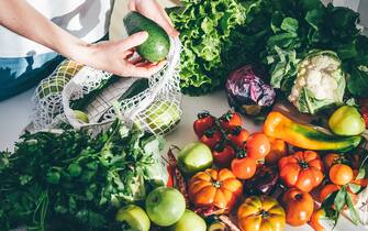 Young woman returned with purchases from grocery store takes fresh organic vegetables out of mesh bag putting on kitchen table at home close view