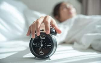 Young asian woman wake up early in morning. Female lying on bed and trying turn off alarm clock. Young sleeping woman and alarm clock in bedroom at home.