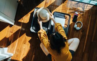 Overhead view of young Asian woman sitting on the floor in her bedroom, packing a suitcase for a trip. Getting ready for a vacation. Traveller's accessories. Travel and vacation concept