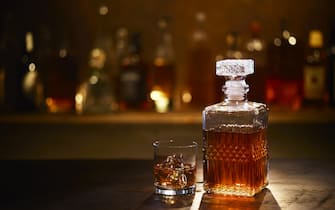 A Bourbon Whiskey Decanter and Bourbon on the Rocks in a Tumbler sitting on a bar with bottles in the background.
