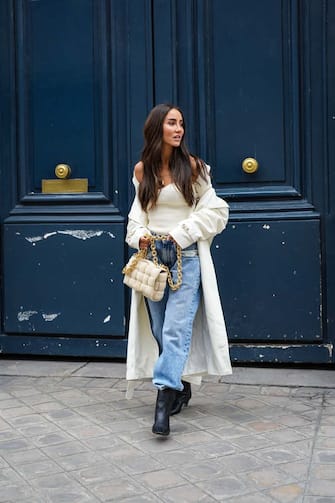 PARIS, FRANCE - APRIL 30: Tamara Kalinic wears gold earrings, a gold chain pendant necklace, a white ribs bustier Khaite top with long sleeves, white long oversized Magda Butrym coat,  blue faded large oversized  Margiela denim jeans pants, a beige suede Bottega Veneta Cassette handbag, black shiny leather pointed heels western Khaite ankle boots, on April 30, 2021 in Paris, France. (Photo by Edward Berthelot/Getty Images)