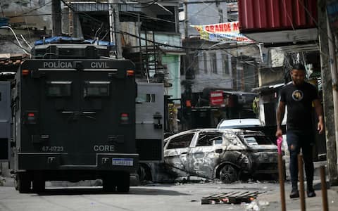 A local walks past a car burnt during a barricade within the Operacao Contencao (Operation Containment) at the Vila Cruzeiro favela, in the Penha complex, in Rio de Janeiro, Brazil, on October 28, 2025. At least 2,500 agents took part in an operation to arrest drug traffickers from the Comando Vermelho (CV), which resulted in, at least, 18 suspects and several police officers dead. (Photo by Mauro PIMENTEL / AFP)