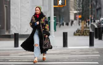 NEW YORK, NEW YORK - FEBRUARY 12: A guest wears a red and green wool hoodie, a black with embroidered multicolored flower pattern long coat with sheep fur borders, a dark brown shiny leather braided zipper handbag, blue faded denim ripped wide legs pants, camel shiny leather with embroidered white flower pattern mules , outside Ulla Johnson, during New York Fashion Week, on February 12, 2023 in New York City. (Photo by Edward Berthelot/Getty Images)