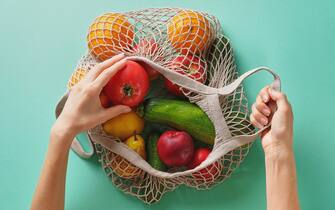 Fresh juicy fruits and vegetables, products in a reusable shopping bag. A girl or woman takes or lays out products from a string bag made from recycled materials on a Green Pastel background. Vegetarianism, Veganism. No plastic.